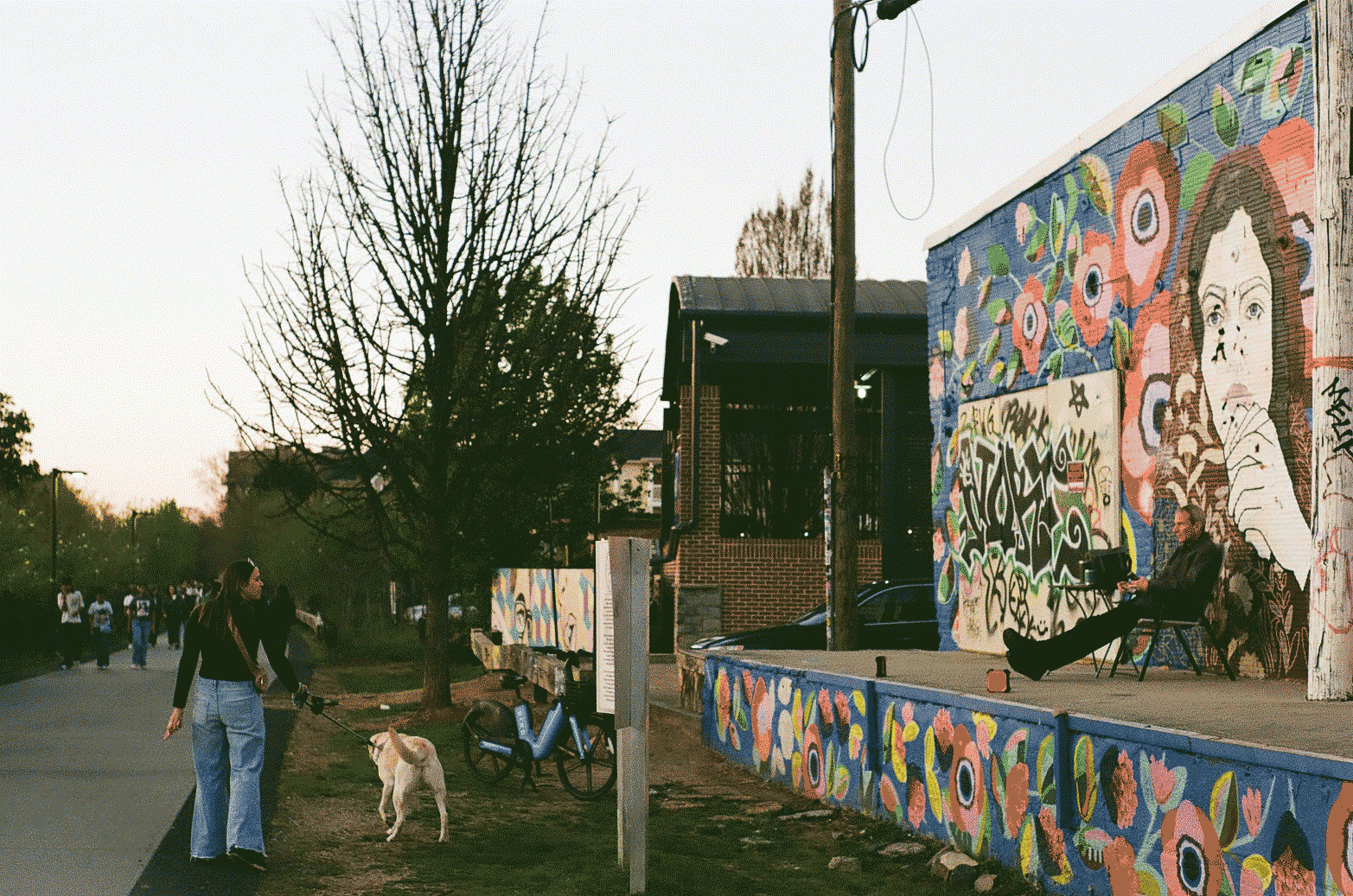 Woman walking a dog along a greenway next to a mural covered building. A man sits relaxed watching them walk by.