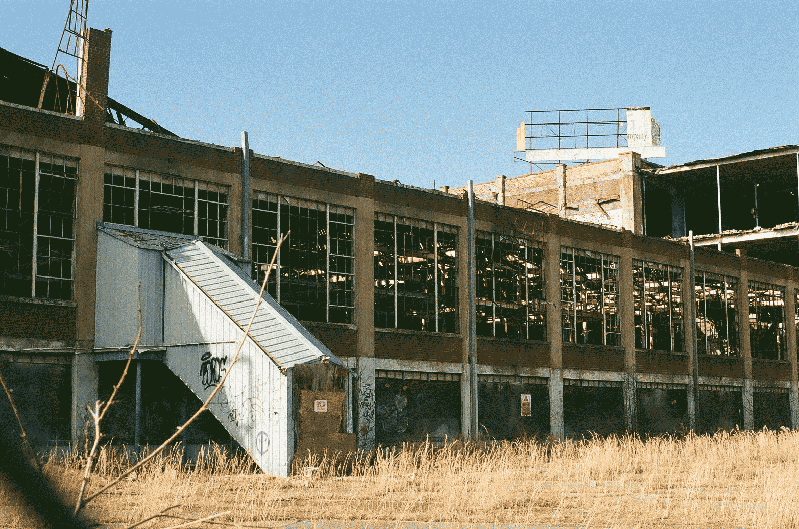 The back of an abandoned factory with windows blown out, cracked sidewalk with tall grass growing